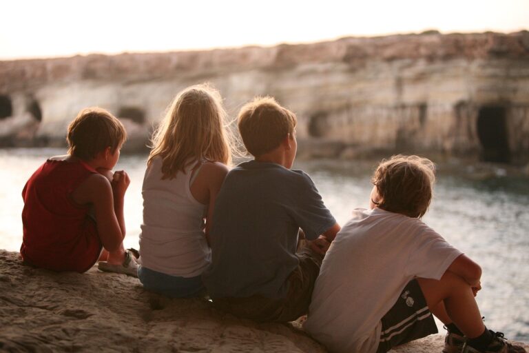 Four children sitting side by side on a rock ledge, viewed from behind, looking out over a body of water with a rocky shoreline in the distance.